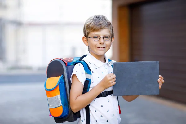 Счастливый маленький мальчик в очках, рюкзаке или сумке. Школьник по дороге в школу. Здоровый очаровательный ребенок на улице с пустым мелом на столе для копирайта. Возвращение в школу . — стоковое фото