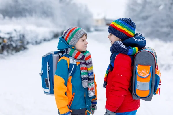 Två små pojkar i grundskolan som går till skolan under snöfallet. Glada barn som har roligt och leker med första snön. Syskon an vän med ryggsäck i färgglada vinterkläder. — Stockfoto