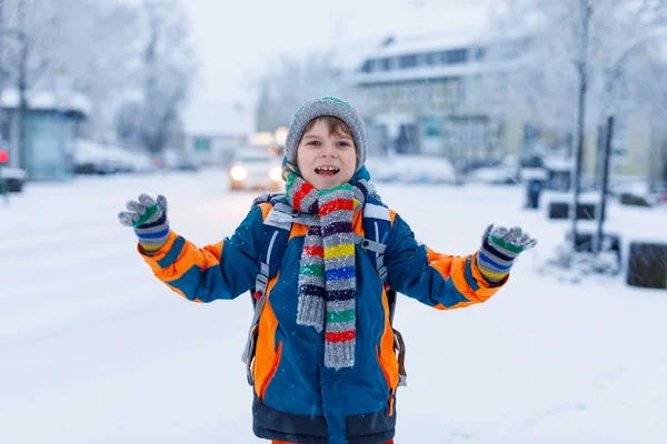 Kleine schooljongen van de lagere klasse die naar school loopt tijdens de sneeuwval. Gelukkig kind dat plezier heeft en speelt met de eerste sneeuw. Leerling met rugzak of tas in kleurrijke winterkleding. — Stockfoto