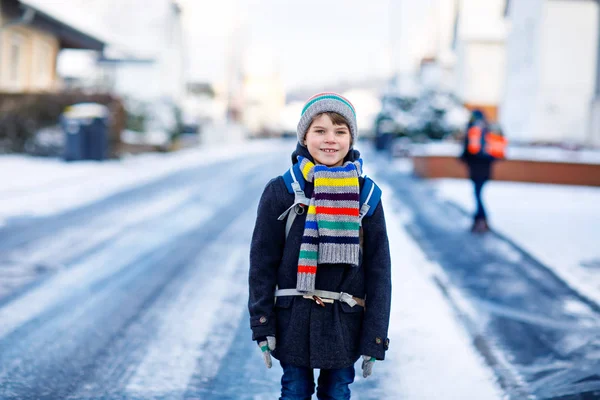 Menino da escola de classe primária a caminhar para a escola durante a queda de neve. Criança feliz se divertindo. Rua da cidade com carros e trânsito. Estudante com mochila ou mochila em roupas coloridas de inverno. — Fotografia de Stock