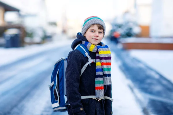 Malý školáček základní třídy, který chodí do školy během sněžení. Šťastné dítě se baví. Ulice s auty a dopravou. Student s batohem nebo brašnou v barevných zimních šatech. — Stock fotografie