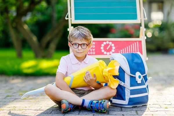 Niño feliz con gafas sentado junto al escritorio y la mochila o mochila. Colegial con cono de bolsa escolar alemán tradicional llamado Schultuete en su primer día en la escuela — Foto de Stock