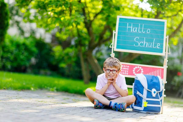 Glad liten unge pojke med glasögon som sitter vid skrivbord och ryggsäck eller axelväska. Schoolkid med traditionella tyska skolväska kallas Schultuete på sin första dag i skolan. Hej skola i tyska. — Stockfoto