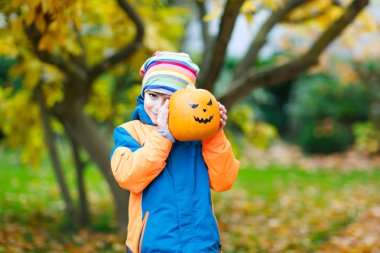 Cadılar bayramı balkabağı feneriyle mutlu küçük çocuk. Renkli elbiseli komik çocuk soğuk sonbahar gününde sonbahar bahçesinde eğleniyor ve oynuyor. Korkunç balkabağı feneri, bayram kutlaması..