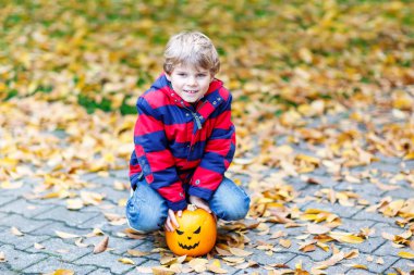 Cadılar bayramı balkabağı feneriyle mutlu küçük çocuk. Renkli elbiseli komik çocuk soğuk sonbahar gününde sonbahar bahçesinde eğleniyor ve oynuyor. Korkunç balkabağı feneri, bayram kutlaması.