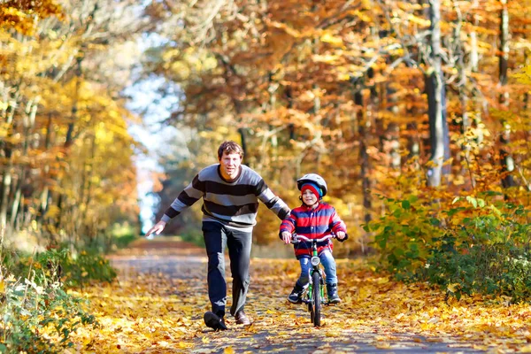 Pequeño niño preescolar y su padre en el parque de otoño con una bicicleta. Papá enseñando a su hijo a andar en bicicleta. Ocio familiar activo. Niño con casco en bicicleta. Seguridad, deportes, ocio con concepto de niños .. — Foto de Stock