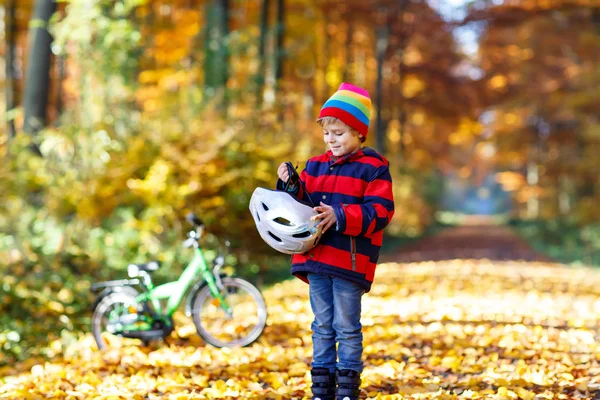 Little kid boy in colorful warm clothes in autumn forest park with a bicycle. Active child putting safe helmet before cycling on sunny fall day in nature. Safety, sports, leisure with kids concept — Stock Photo, Image