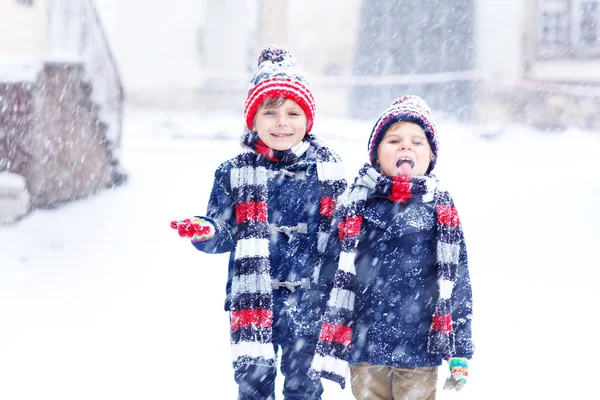 Renkli kıyafetleri olan iki küçük çocuk şiddetli kar yağışı sırasında dışarıda oynuyorlar. Soğuk günlerde kışın çocuklarla aktif bir eğlence. Mutlu kardeşler ve ikizler ilk karla eğleniyorlar.. — Stok fotoğraf