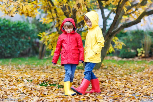 Dos mejores amigos y los niños chicos otoño parque en ropa colorida. Niños felices hermanos divirtiéndose en capas de lluvia rojo y amarillo y botas de goma. Familia jugando al aire libre. ocio activo. — Foto de Stock