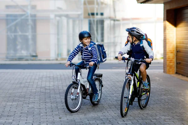 Dva školáci v bezpečnostní helmě na kole ve městě s batohy. Šťastné děti v barevných šatech na kolech na cestě do školy. Bezpečný způsob pro děti venku do školy — Stock fotografie