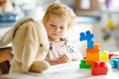 Bebek odasında eğitici oyuncaklarla oynayan pelüş tavşanlı sevimli bir kız. Mutlu sağlıklı çocuk evde renkli farklı plastik bloklarla eğleniyor. Sevimli bebek öğrenimi yaratma.