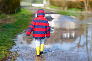 Sarı yağmur giyen küçük çocuk çocuk çizme ve yürüme ve içine atlama bahar günü sıcak güneşli birikintisi.