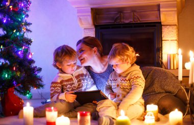 Genç anne ve Noel zamanında bir şöminenin evde oturan iki küçük çocuğu. Tablet ile oynayan çocuklar çocuklar. Üç sarılma aile. Mutlu kadın ve sevimli oğlu, İkizler küçük çocuklar