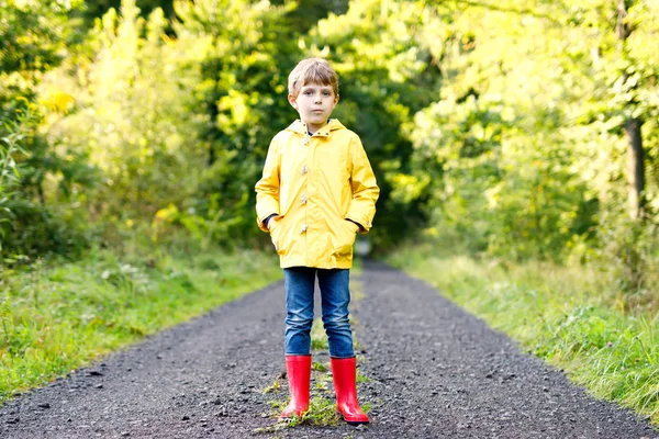 Retrato de hermoso niño sonriente en chaqueta de lluvia amarilla y botas de goma roja. Niño feliz contra el fondo verde del árbol. Niño jugando al aire libre —  Fotos de Stock