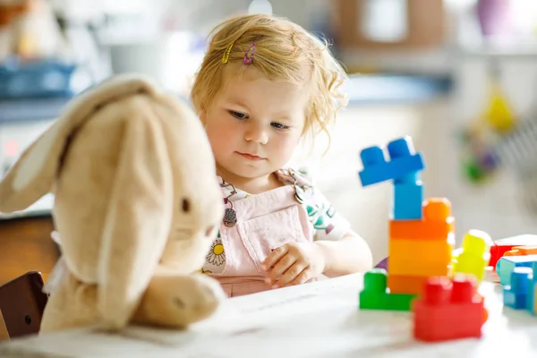 Adorable niña con conejito de peluche favorito jugando con juguetes educativos en la guardería. Feliz niño sano que se divierte con diferentes bloques de plástico de colores en casa. Lindo bebé aprendizaje creación. — Foto de Stock