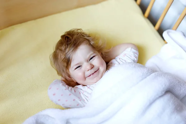 寝る前にかわいい赤ん坊の女の子がベッドに寝そべっている。ベッドで幸せな穏やかな子供。眠りにつく。親を見ている平和的で笑顔の子供。一人で寝る — ストック写真