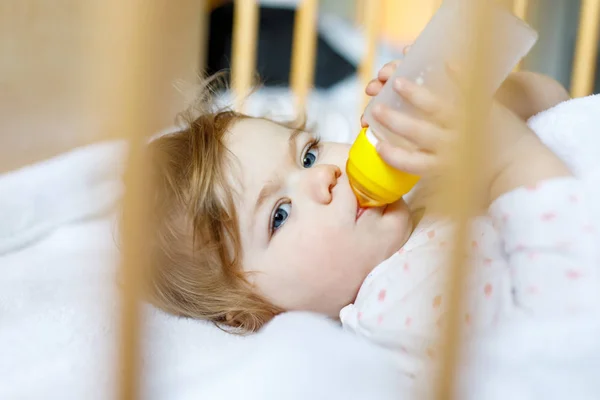 Милая маленькая девочка держит бутылочку с формулой мягкой и питьевой. Ребенок в детской кроватке перед сном — стоковое фото