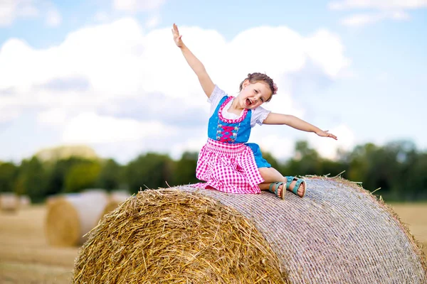 Roztomilá holčička v tradičním bavorském kostýmu na pšeničném poli. Německé dítě s balíkem sena během Oktoberfestu v Mnichově. Předškolní dívka hrát na balíky sena během letní sklizně v Německu. — Stock fotografie