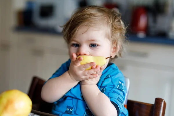 Linda adorable niña comiendo pera fresca. Bebé feliz hambriento de un año sosteniendo fruta. Chica en la cocina doméstica, teniendo bocadillos de comida saludable. Niño rubio sonriente — Foto de Stock