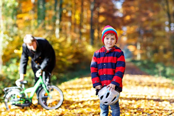 Little kid boy of five years and his father in autumn forest with a bicycle. Active child putting his bike helmet. Safety, sports, leisure with kids concept. Man with bike on background. — Stock Photo, Image