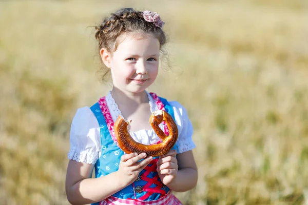 Cute little kid girl in traditional Bavarian costume in wheat field. German child with hay bale during Oktoberfest in Munich. Preschool girl eating pretzel during harvest time in Germany. — Stock Photo, Image