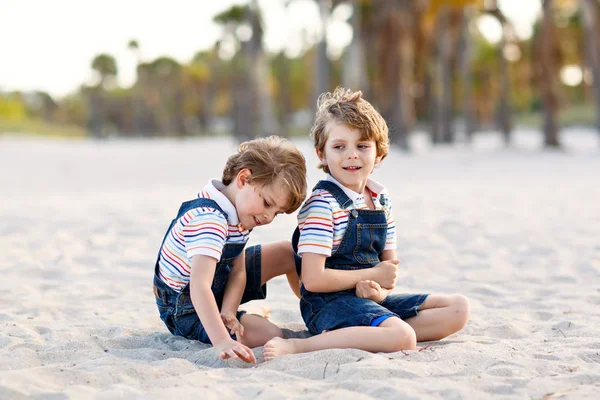 Due adorabili fratellini seduti sulla sabbia sulla spiaggia tropicale, migliori amici felici, ragazzi che giocano, concetto di amicizia. Fratelli fratelli, i gemelli in famiglia guardano con palme. Vacanze in famiglia. — Foto Stock