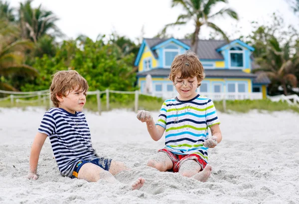 Dwa małe dzieci chłopcy zabawy z budynku zamku z piasku na tropikalnej plaży, szczęśliwy przyjaciele gry. Rodzeństwo braci, bliźniaki z palmami i dom na tle. Życie rodzinne w Los Angeles — Zdjęcie stockowe