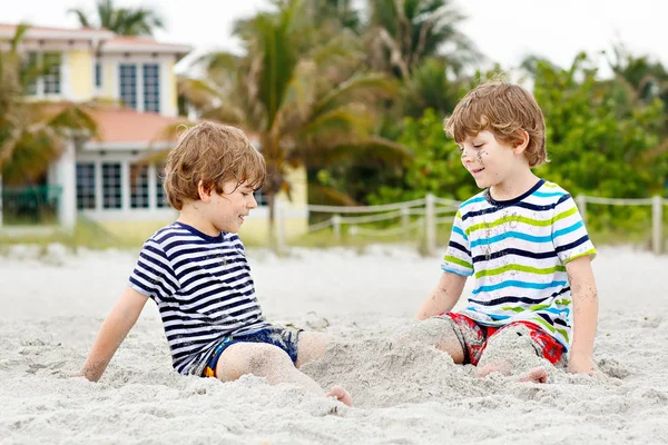Two little kids boys having fun with building sand castle on tropical beach, happy best friends playing. Siblings brothers, twins with palms and home on background. Family life in Los Angeles — Stock Photo, Image