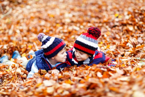 Dois meninos gêmeos jazendo em folhas de outono em roupas coloridas. Feliz irmãos crianças se divertindo na floresta de outono ou parque no dia de outono. Com chapéus de moda casuais e lenços. Amigos brincando juntos. — Fotografia de Stock