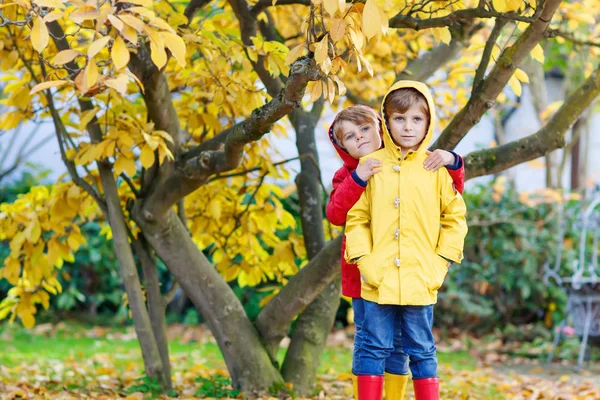 Two little best friends and kids boys autumn park in colorful clothes. Happy siblings children having fun in red and yellow rain coats and rubber boots. Family playing outdoors. active leisure. — Stock Photo, Image