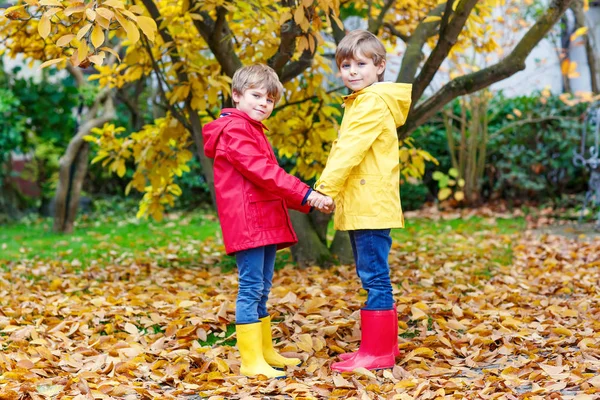 Dos pequeños mejores amigos y niños parque de otoño en ropa de colores. Felices hermanos niños divirtiéndose en abrigos de lluvia rojos y amarillos y botas de goma. Familia jugando al aire libre. ocio activo. — Foto de Stock