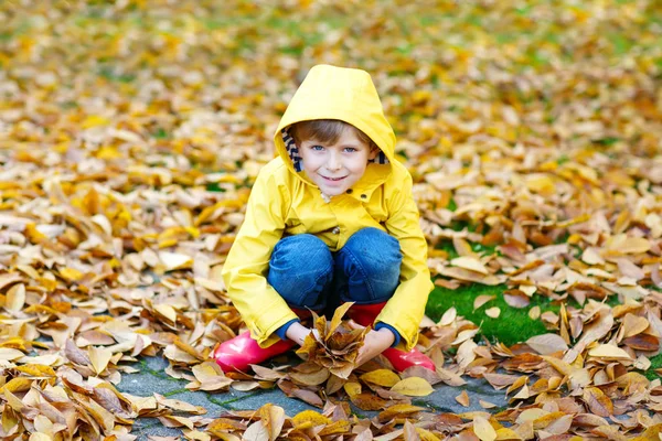 Retrato de niño lindo feliz en abrigo de lluvia amarillo y botas de goma roja con fondo de hojas de otoño. Divertido niño divirtiéndose y jugando en bosque de otoño o parque en el frío día otoñal — Foto de Stock