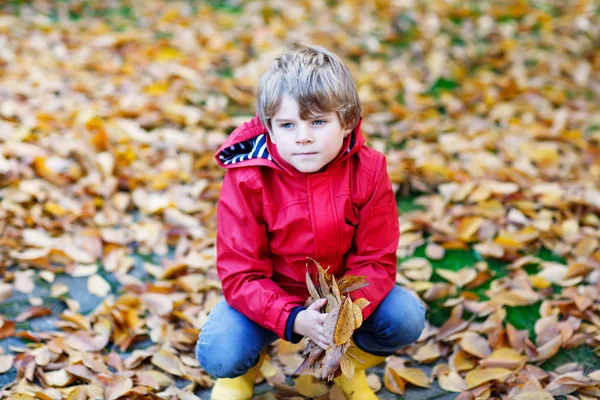 Portrait of happy cute little kid boy in red rain coat and yellow rubber boots with autumn leaves background. Funny child having fun and playing in fall forest or park on cold autumnal day — Stock Photo, Image