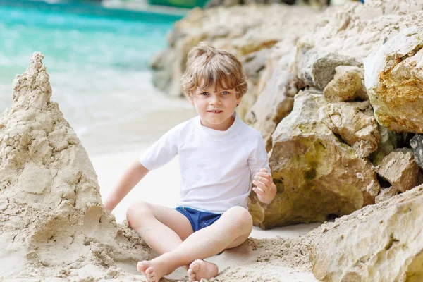 Happy funny little preschool kid boy having fun with building a sand castle on ocean beach. child playing on family vacations on tropical island. summer, happiness, childhood concept. — Stock Photo, Image