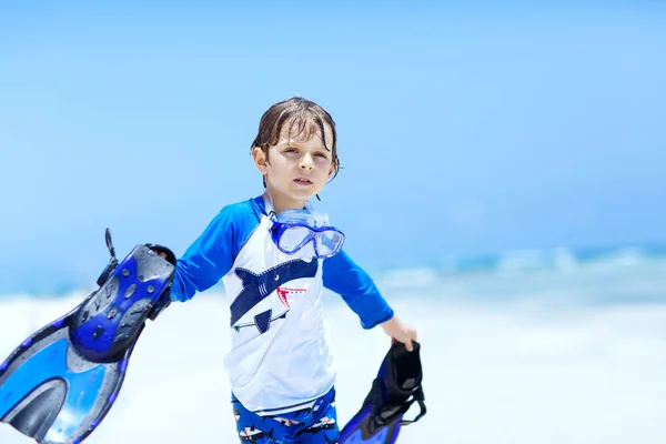 Adorable little blond kid boy having fun on tropical beach of Maldives. Excited child playing and surfing in sun protected swimsuit in ocean on vacations. White sand, Kid holding flippers for swimming — Stock Photo, Image