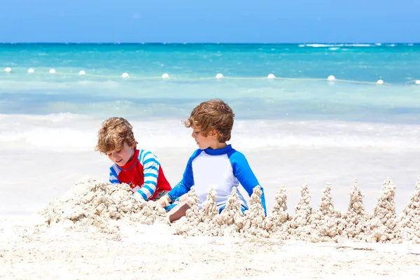 İki küçük çocuk Meksika Playa del Carmen plajında kumdan kale inşa ederken eğleniyor. Çocuklar tatillerinde birlikte oynuyorlar. İkizler, mutlu kardeşler gülüyor ve gülüyorlar.. — Stok fotoğraf