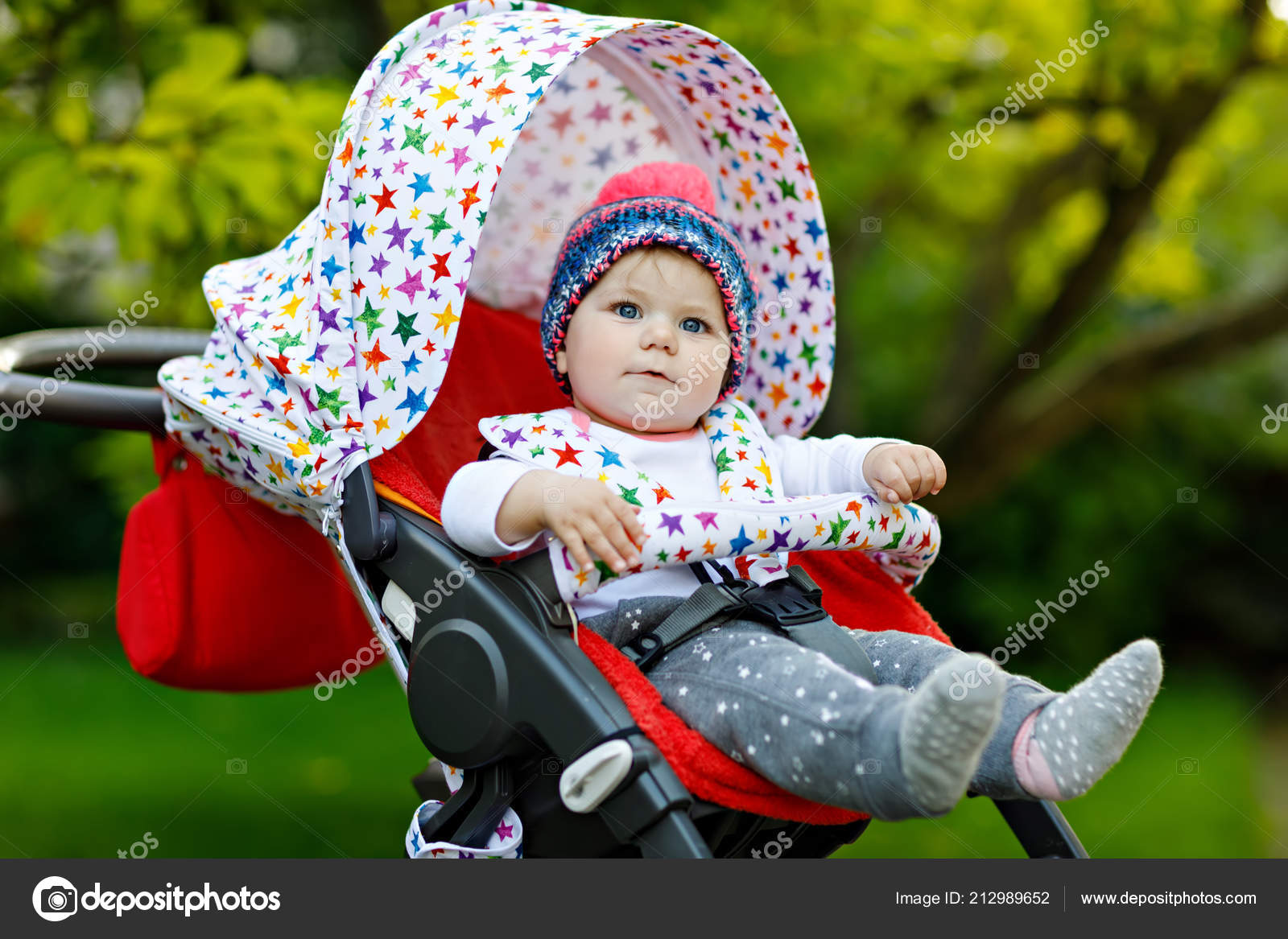 Cute little beautiful baby girl sitting in the pram or stroller and waiting  for mom. Happy smiling child with blue eyes. With green tree background.  Baby daughter going for a walk Stock