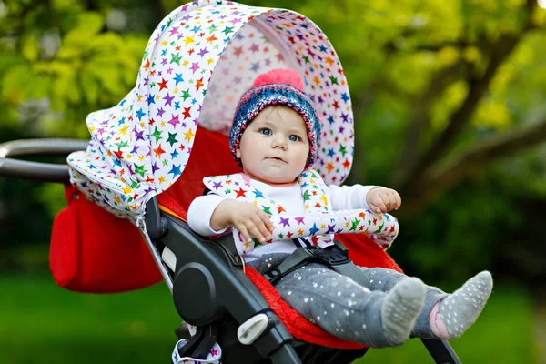 Bonito menina bonita sentada no carrinho de bebê ou carrinho e esperando a mãe. Criança sorridente feliz com olhos azuis. Com fundo de árvore verde. Filha bebé vai dar um passeio — Fotografia de Stock