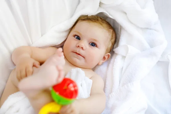 Niedliches kleines Baby, das nach dem Baden mit Spielzeugrassel und eigenen Füßen spielt. entzückende schöne Mädchen in weiße Handtücher gehüllt — Stockfoto