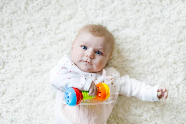 Leuke schattige pasgeboren baby spelen met kleurrijke rammelaar speelgoed op witte achtergrond. Nieuw geboren kindje, kleine meisje te kijken naar de camera. Familie, nieuw leven, kindertijd, begin concept. Baby leren pak. — Stockfoto