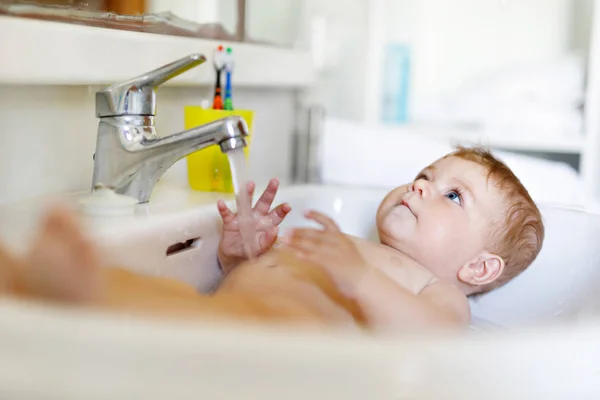 Schattige schattige baby nemen bad in wastafel en pak waterkraan. Klein gezond meisje met grote blauwe ogen die plezier hebben. Schoon mooi kind spelen — Stockfoto