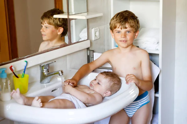 Schattige schattige baby nemen bad in wastafel en pak waterkraan. Jongetje helpt en speelt met zusje. Gelukkige broer en kind hebben plezier. Gezonde kinderen samen in de badkamer. Gezinstijd. — Stockfoto
