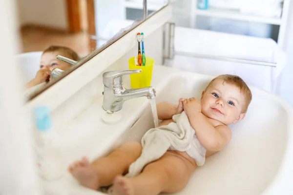 Cute adorable baby taking bath in washing sink and grab water tap. Little healthy girl with big blue eyes having fun. Clean beautiful child playing — Stock Photo, Image