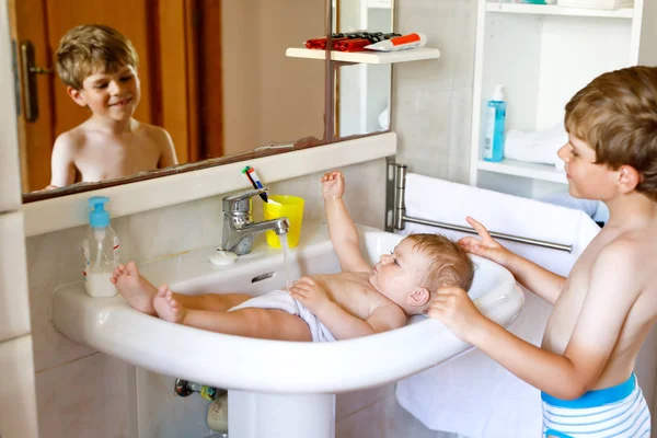 洗面台でお風呂に入り、水汲みをしている可愛い赤ちゃん。子供の男の子は妹と一緒に助けて遊んでいます。幸せな兄弟と子供は楽しんでいます。一緒に浴室で健康な子供たち。家族の時間. — ストック写真