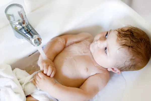 Schattige schattige baby nemen bad in wastafel en pak waterkraan. Klein gezond meisje met grote blauwe ogen die plezier hebben. Schoon mooi kind spelen — Stockfoto