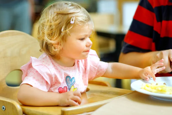 Αξιολάτρευτο μικρό παιδί κορίτσι τρώει υγιεινά λαχανικά και ανθυγιεινά τηγανιτές πατάτες. Χαριτωμένο μωρό ευτυχισμένο παιδί παίρνοντας τροφή από γονείς πιάτο σε εστιατόριο — Φωτογραφία Αρχείου