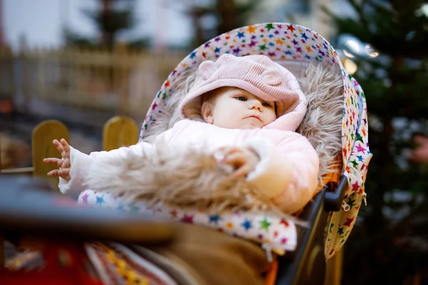 Aranyos kis gyönyörű kislány ül a babakocsiban, vagy babakocsi télen este a karácsonyi piacon. Boldog mosolygós gyermek meleg ruhákban, divat stílusos baba általában — Stock Fotó