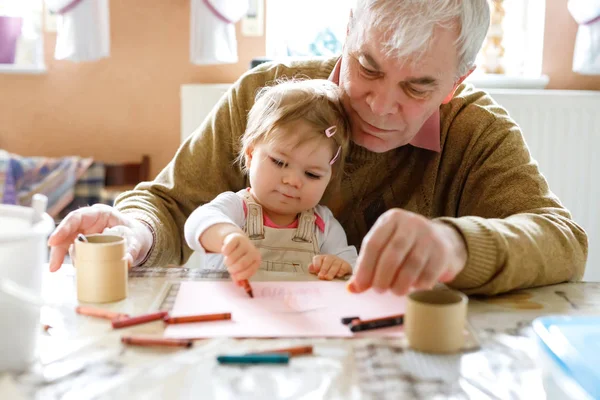 Menina pequena bonito bebê criança e bonito avô sênior pintura com lápis coloridos em casa. Neto e homem se divertindo juntos. Família e geração apaixonadas — Fotografia de Stock
