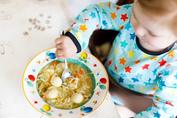 Söt liten flicka som äter från sked grönsaksnudelsoppa. Livsmedel, barn, foder och utvecklingskoncept. Söt småbarn, dotter med sked sittande i barnstol och lär sig att äta själv. — Stockfoto