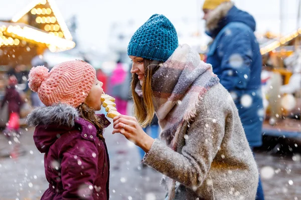 Young mother and daughter eating white chocolate covered fruits on skewer on traditional German Christmas market. Happy girl and woman on traditional family market in Germany, Munich during snowy day. — Stock Photo, Image